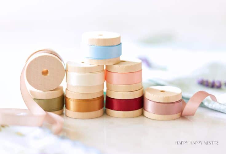 A collection of colorful ribbon spools stacked in a neat arrangement on a light surface. The ribbons include shades of blue, pink, red, olive, and beige. The setup has a soft, pastel aesthetic with a blurred background, giving a cozy and creative vibe.