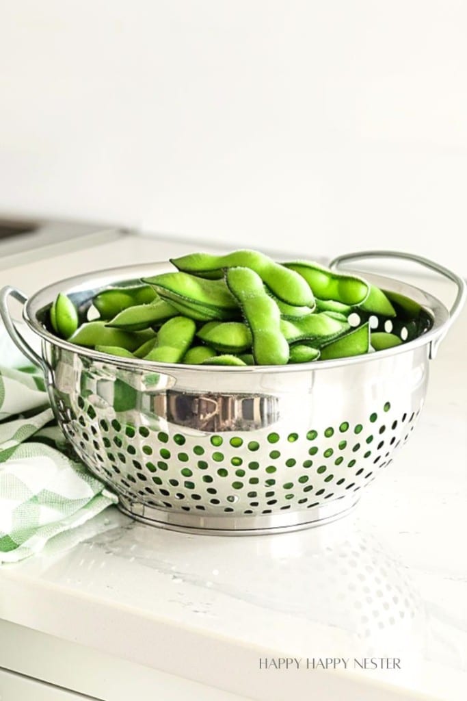 A stainless steel colander is filled with fresh, green edamame pods. The colander features circular holes and handles on each side, and it is placed on a white countertop. A green and white checkered cloth is partially visible next to the colander.