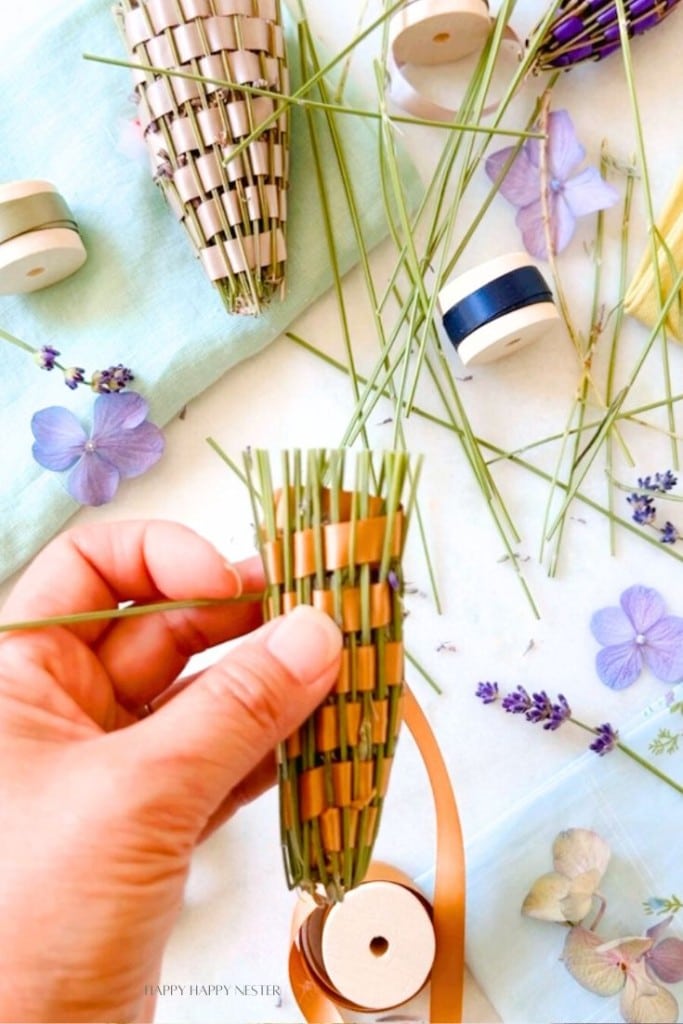 A hand holds a small woven cone made of dried grass stems. Spools of ribbon, dried lavender, and pressed flowers are scattered on a light blue fabric in the background. The crafting materials suggest a DIY floral arrangement or decorative project in progress.