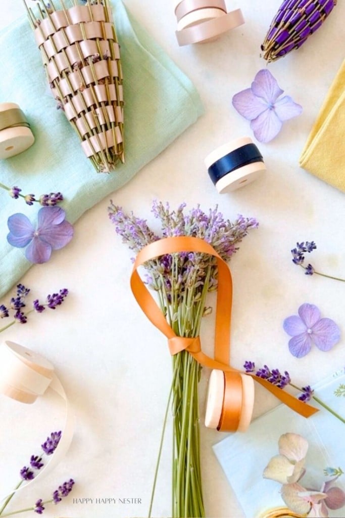 A bouquet of lavender tied with an orange ribbon lies on a white surface surrounded by lavender sprigs, purple flowers, ribbons, and craft supplies. The scene is bright and neatly arranged, evoking a sense of creativity and calm.