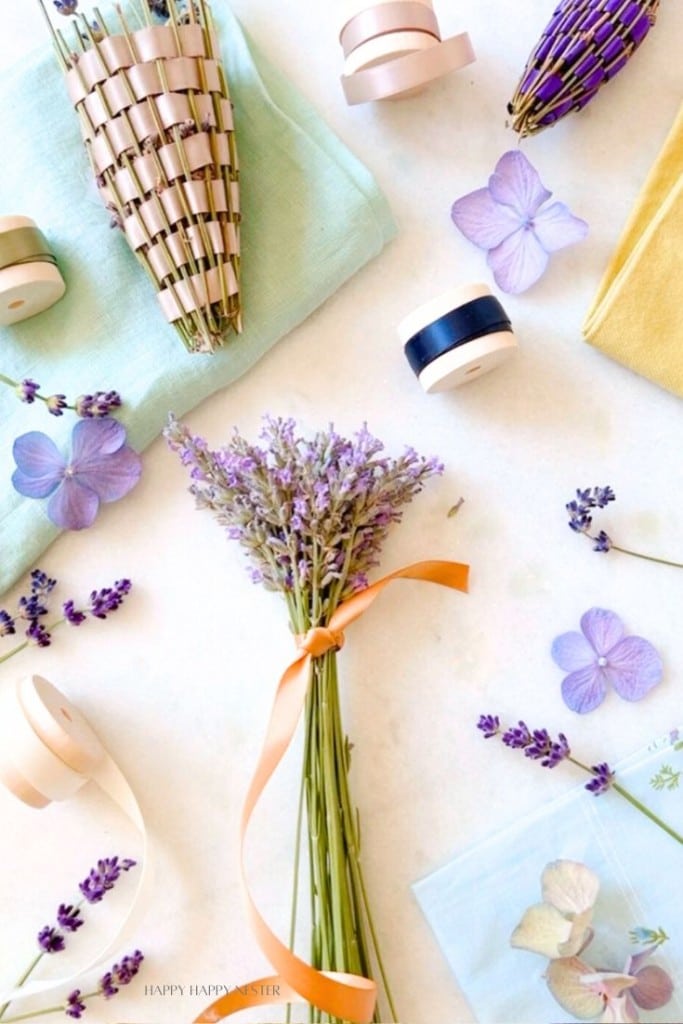 A crafting scene with items for making lavender sachets. A bunch of dried lavender tied with an orange ribbon is in the center, surrounded by rolls of ribbon, woven cones, lavender sprigs, and blue and purple flower petals on a light surface.