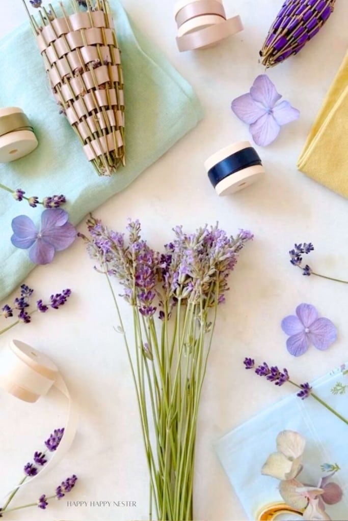 A collection of crafting supplies scattered on a white surface, including bundles of dried lavender, purple hydrangea petals, spools of ribbon, paper cones, and pastel-colored fabrics. The scene exudes a calm and creative atmosphere.