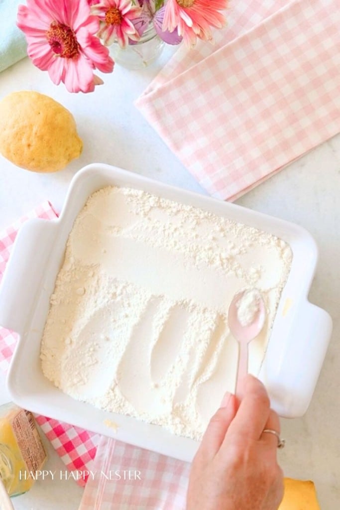 A white baking dish filled with flour is being smoothed out by a hand holding a spoon. Nearby, there are lemons, a pink and white checkered cloth, a pink floral napkin, and a small bouquet of pink flowers in a jar.