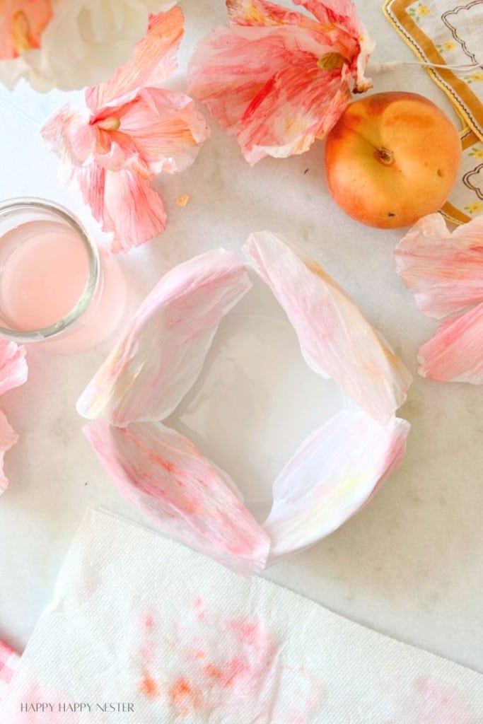 An arrangement of pink flower petals surrounds a white dish on a marble surface. Next to the dish is a peach, a glass of pink liquid, and a paper towel with pink stains. The setting appears to be delicate and artistic.