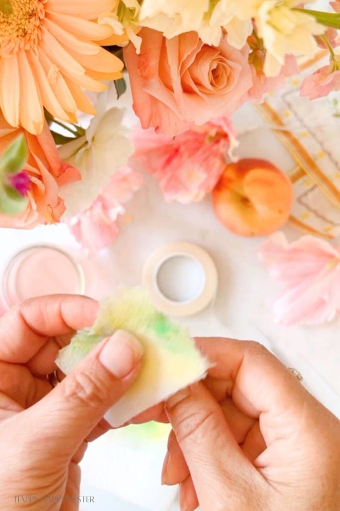 Hands holding a piece of craft material, possibly a paper flower, with various colorful flowers, a peach, and crafting supplies like a roll of tape visible in the background. The scene is bright and appears to be set up for a floral crafting project.