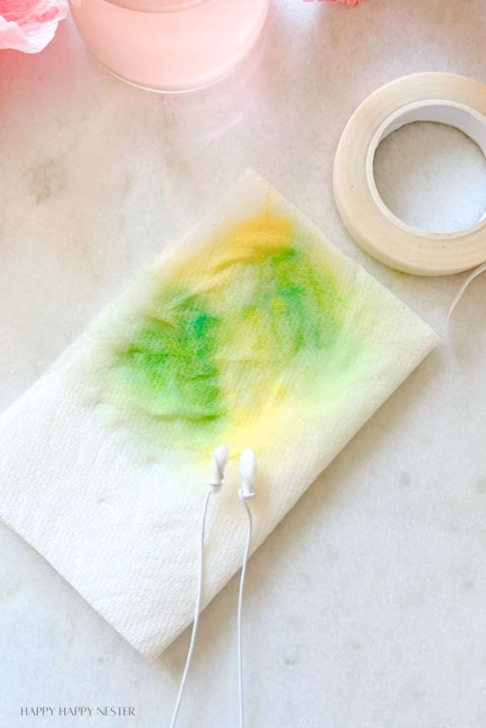 A pair of white earphones is placed on a white paper towel, which has been stained with vibrant green and yellow ink. Next to the paper towel is a roll of white tape. The background is a light, marble-textured surface.