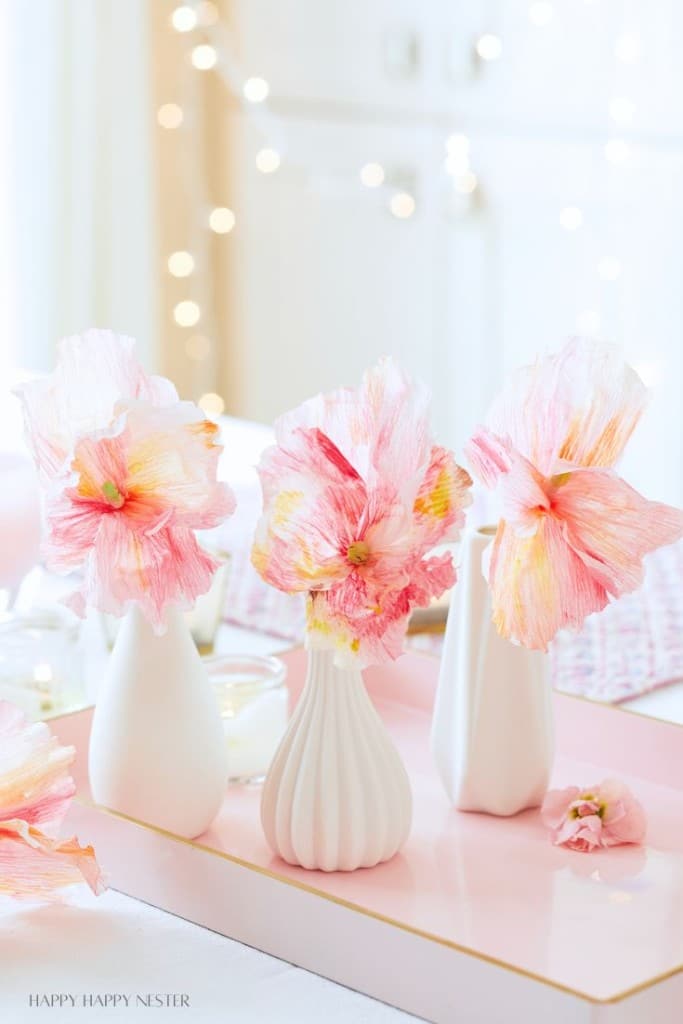 Three delicate pink and white crepe paper flowers are arranged in white vases on a table. A string of warm, glowing fairy lights softly illuminates the background, creating a cozy and elegant atmosphere. A single crepe flower petal lies nearby.