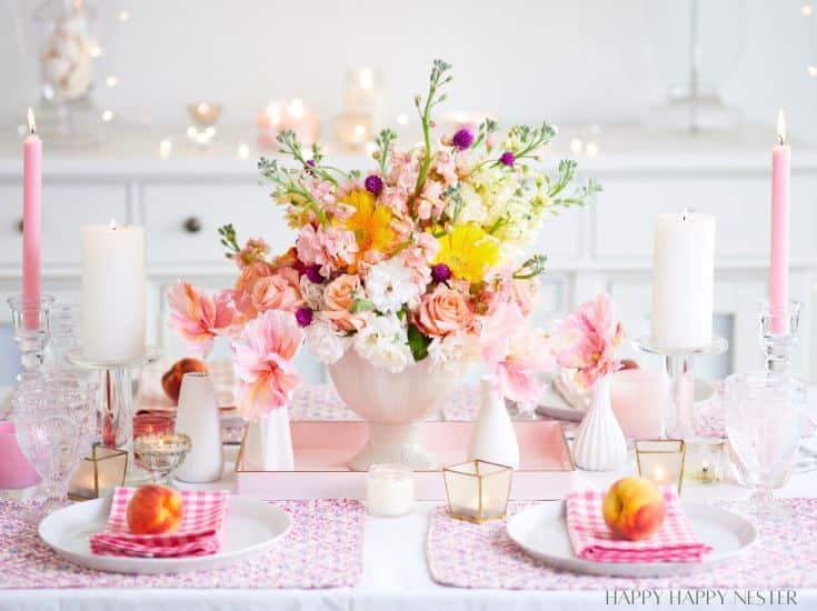 A beautifully set dining table featuring a centerpiece with an arrangement of pink, white, and yellow flowers. The table has white plates with pink checkered napkins, candles in glass holders, and peaches on either side. Twinkling fairy lights adorn the background.