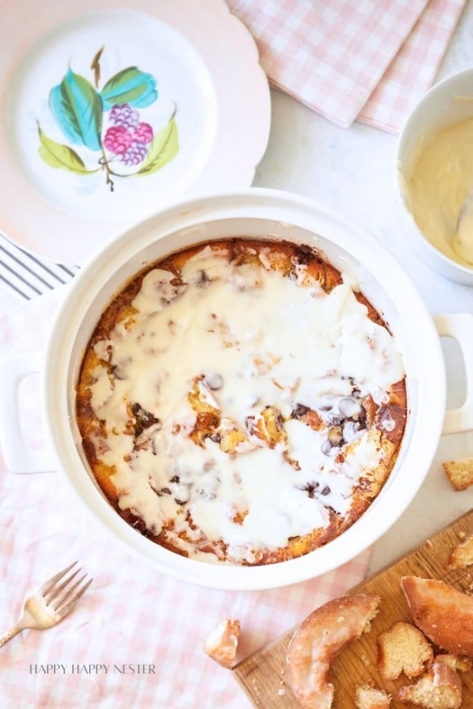 A baked dessert in a round white dish is covered with a white glaze. It rests on a table with a pink checkered cloth, near a plate with floral design and a white bowl containing more glaze. Some dessert pieces and forks are scattered around.