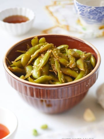 A brown bowl filled with seasoned edamame sits on a table surrounded by a teacup, a small sauce dish, and a printed napkin. The background is softly blurred, emphasizing the edamame and suggesting a casual dining setup.