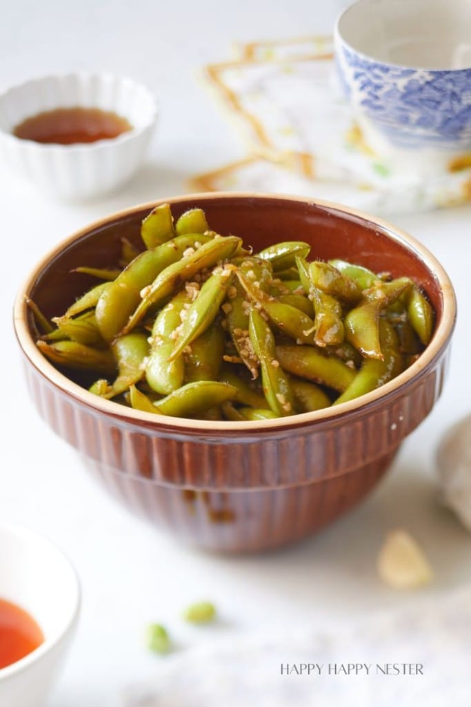 A brown bowl filled with seasoned edamame sits on a table surrounded by a teacup, a small sauce dish, and a printed napkin. The background is softly blurred, emphasizing the edamame and suggesting a casual dining setup.