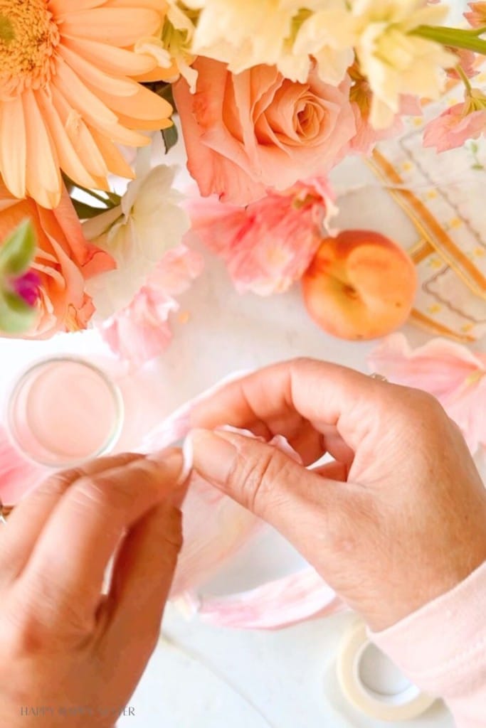 Close-up of hands crafting a flower-shaped pom-pom with delicate white and pink material. The background features a vibrant arrangement of fresh flowers in various colors and an apricot, creating a bright and cheerful atmosphere.