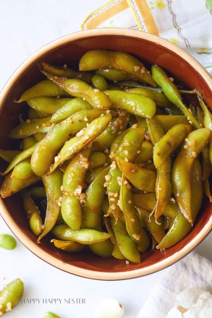 A close-up of a bowl filled with cooked edamame pods, seasoned with garlic and small visible bits of seasoning. The dish is placed on a white surface with part of a patterned cloth napkin and a few scattered edamame pods nearby.