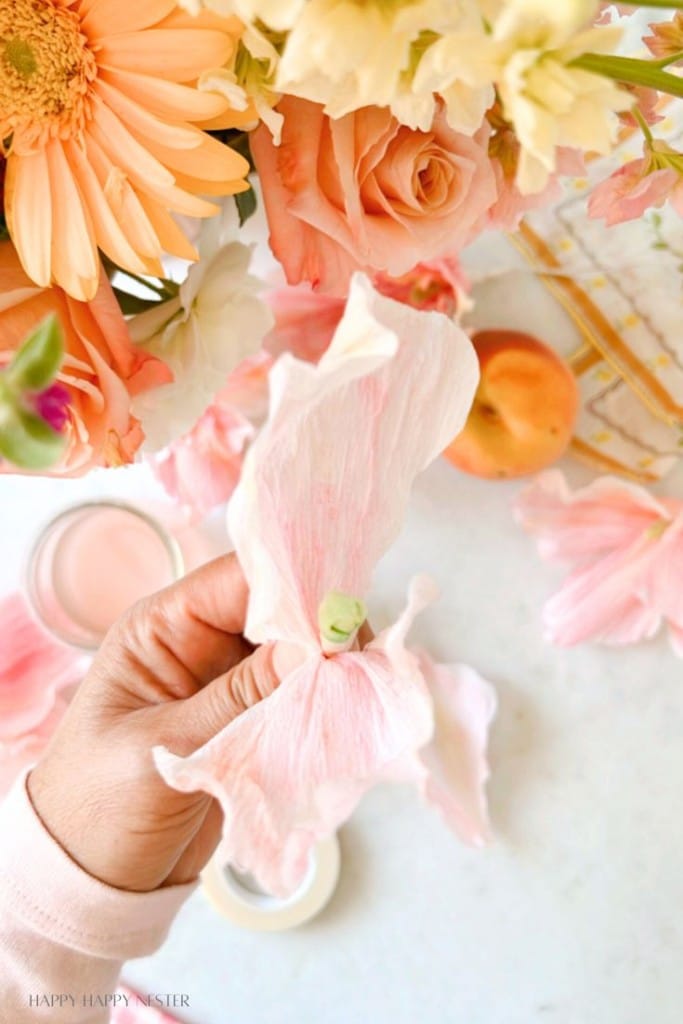 A close-up of a hand holding a delicate pink and white flower petal. Surrounding the hand are various flowers, including roses and gerbera daisies, in soft pastel colors. An apricot and a pair of glasses are also visible on the light-colored surface.