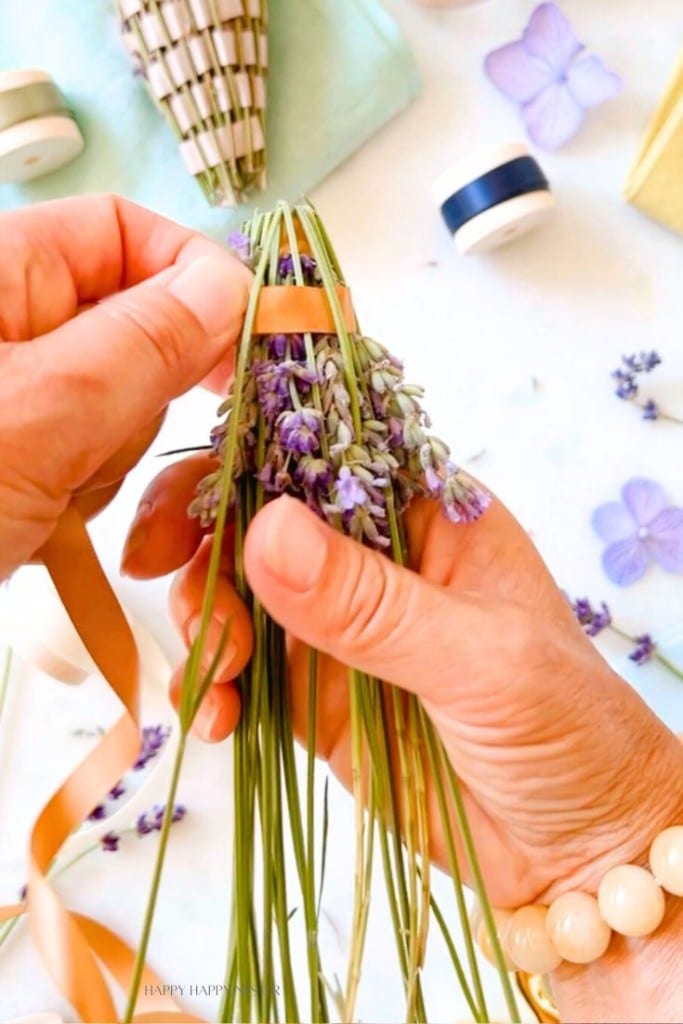 A pair of hands is weaving a lavender wand with a ribbon. Fresh lavender stems with small purple flowers and green leaves are being wrapped tightly together. Craft supplies, such as ribbons and additional lavender stems, are visible in the background.