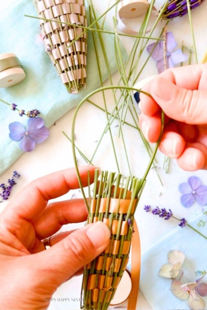 Two hands are shown weaving a lavender wand using fresh lavender stems and ribbon. The partially completed wand lies on a light blue cloth, surrounded by other supplies including more lavender stems, ribbon spools, and scattered flower petals.