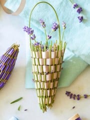 A woven cone basket filled with fresh lavender sprigs lies on a light blue cloth. The basket is woven with green stems and beige ribbon, resembling a cornucopia. Additional sprigs and a purple-wrapped lavender bundle rest nearby on a white surface.