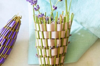A woven cone basket filled with fresh lavender sprigs lies on a light blue cloth. The basket is woven with green stems and beige ribbon, resembling a cornucopia. Additional sprigs and a purple-wrapped lavender bundle rest nearby on a white surface.