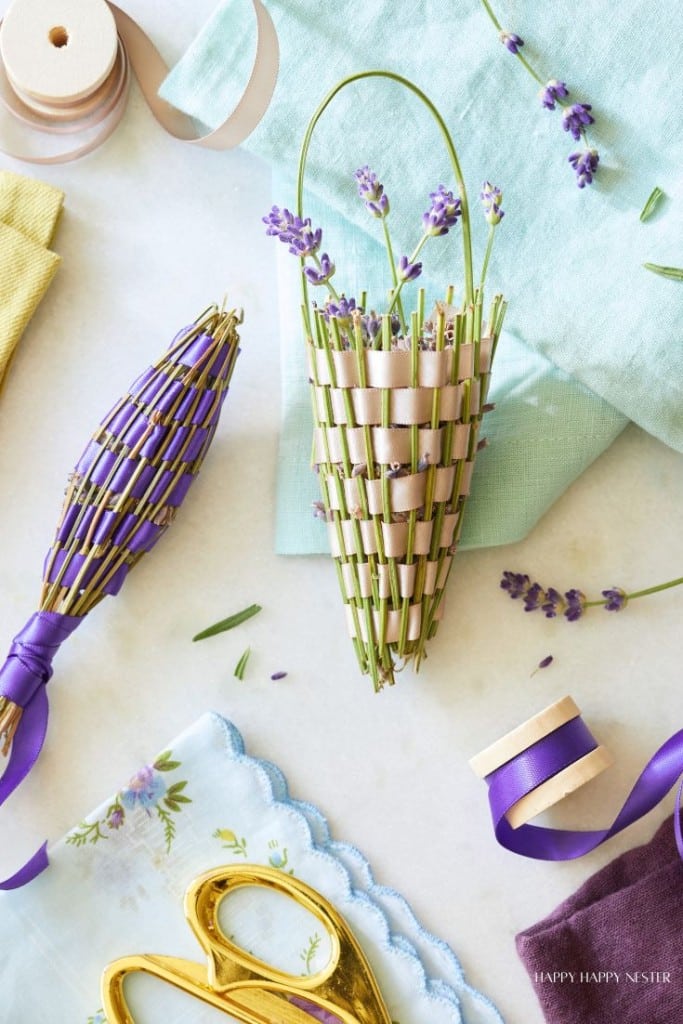 A flat lay of crafting materials featuring a woven lavender bouquet on a light blue cloth. Surrounding items include another lavender bouquet tied with purple ribbon, scissors, ribbon spools, a yellow cloth, and a small floral handkerchief—perfect for your next lavender wands tutorial.