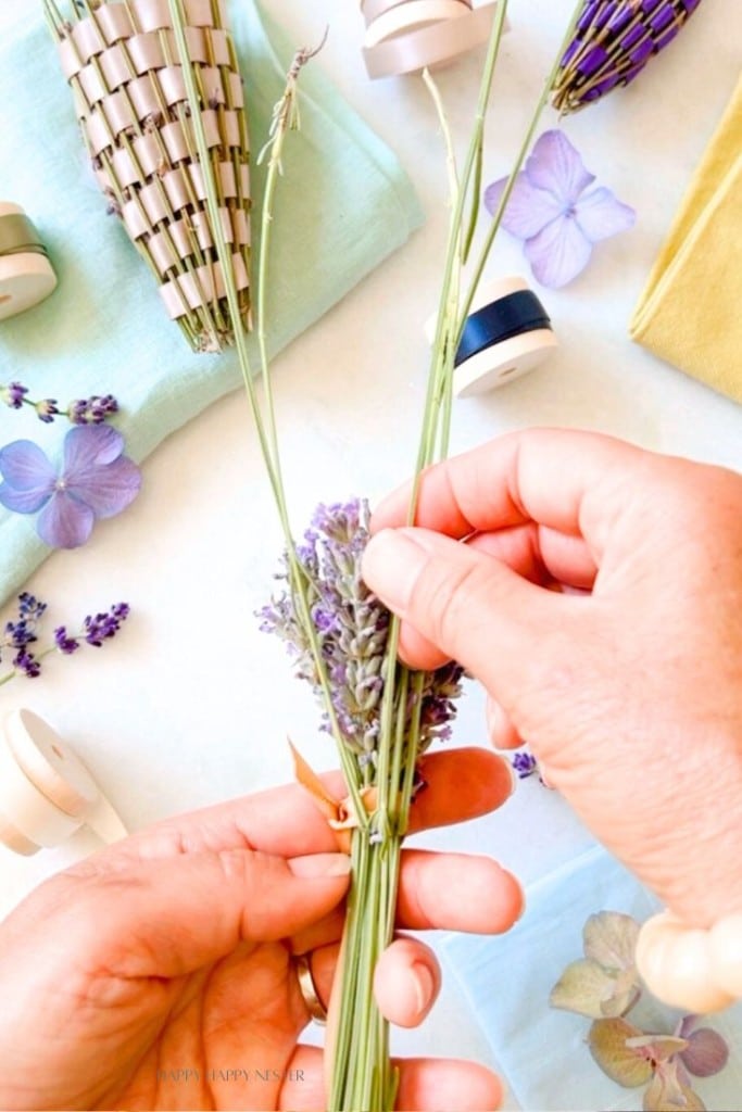 Two hands are crafting, weaving together stems of lavender with thin ribbons. Various crafting supplies, including ribbons, lavender stems, and dried flowers, are scattered on the table. The scene is brightly lit and colorful, highlighting the crafting activity.