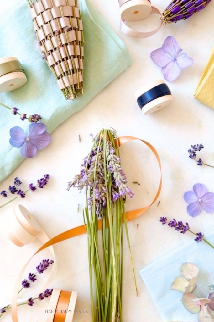 A flat lay of a crafting setup features fresh lavender bunches, spools of ribbon, and pressed flower petals arranged on a light surface. Craft tools and materials, including a small woven basket and fabric, are scattered around, creating a visually pleasing assembly.