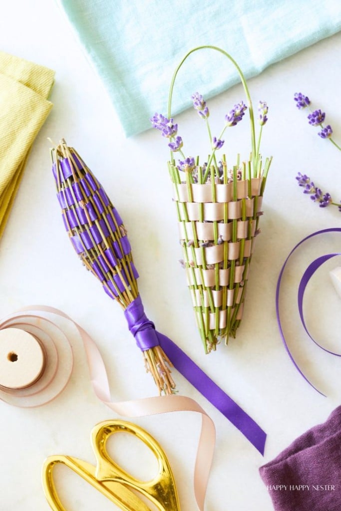 An overhead view of two lavender sachets woven with purple ribbon. One is tied with a purple ribbon, while the other has a pink ribbon. Beside them are crafting supplies including ribbon, gold scissors, and folded colorful fabrics.