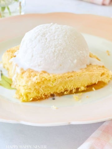 A slice of yellow cake topped with a scoop of vanilla ice cream is served on a pastel-colored plate. In the background, a pink and white striped napkin and a glass vase with flowers are partially visible. The text "HAPPY HAPPY NESTER" appears at the bottom.