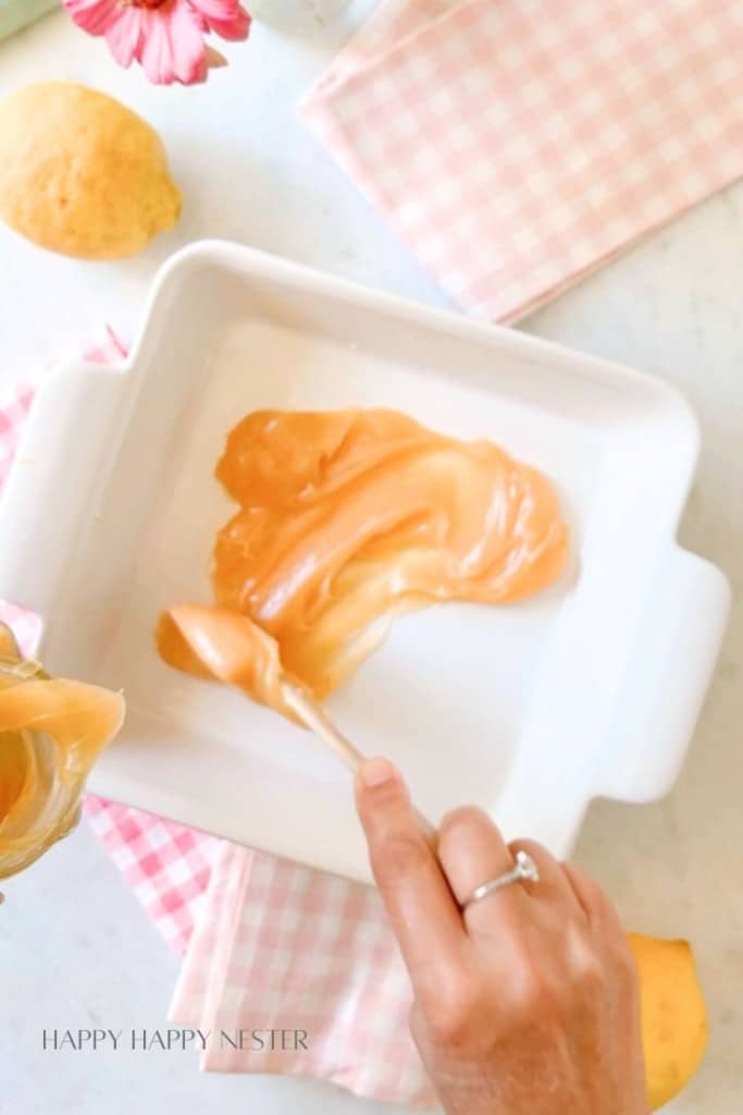 A hand spreading a layer of caramel sauce inside a white square baking dish. Nearby are a lemon, a pink checkered cloth, and a pink flower partially visible at the top. The scene is bright and cheerful. The words "Happy Happy Nester" are visible at the bottom left.