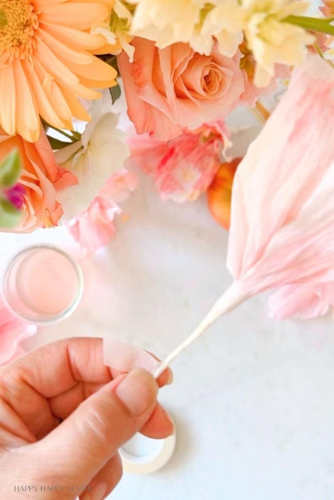 A hand holding a delicate pink flower petal in front of a vibrant bouquet of fresh flowers including roses, gerbera daisies, and other blossoms. The background includes a small glass jar and a pastel-colored flat surface.