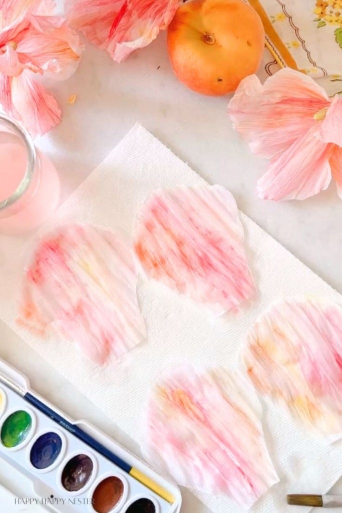 Various shades of peach-colored petals lie on a white paper towel next to a watercolor paint set, a small glass of pink water, and a whole peach. Bright, pastel hues decorate the scene, suggesting a creative art project involving watercolors and craft petals.