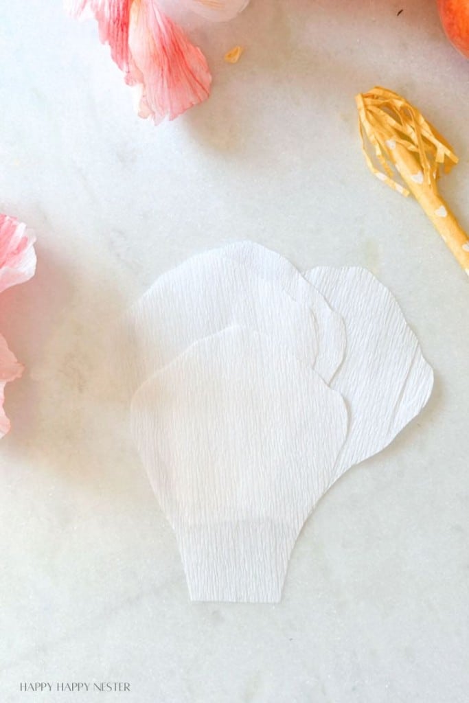 Three white, crepe paper petals are arranged on a light-colored surface. Surrounding the petals are additional crafting supplies, including pink crepe paper and a yellow tool. The setup suggests a crafting project, likely involving floral designs.