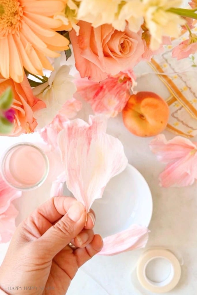 A person's hand holding a pink and white flower petal over a table with various pink and white flowers, an apricot, a small glass of pink liquid, and a white plate. The scene is bright and colorful, suggestive of a delicate, summery atmosphere.