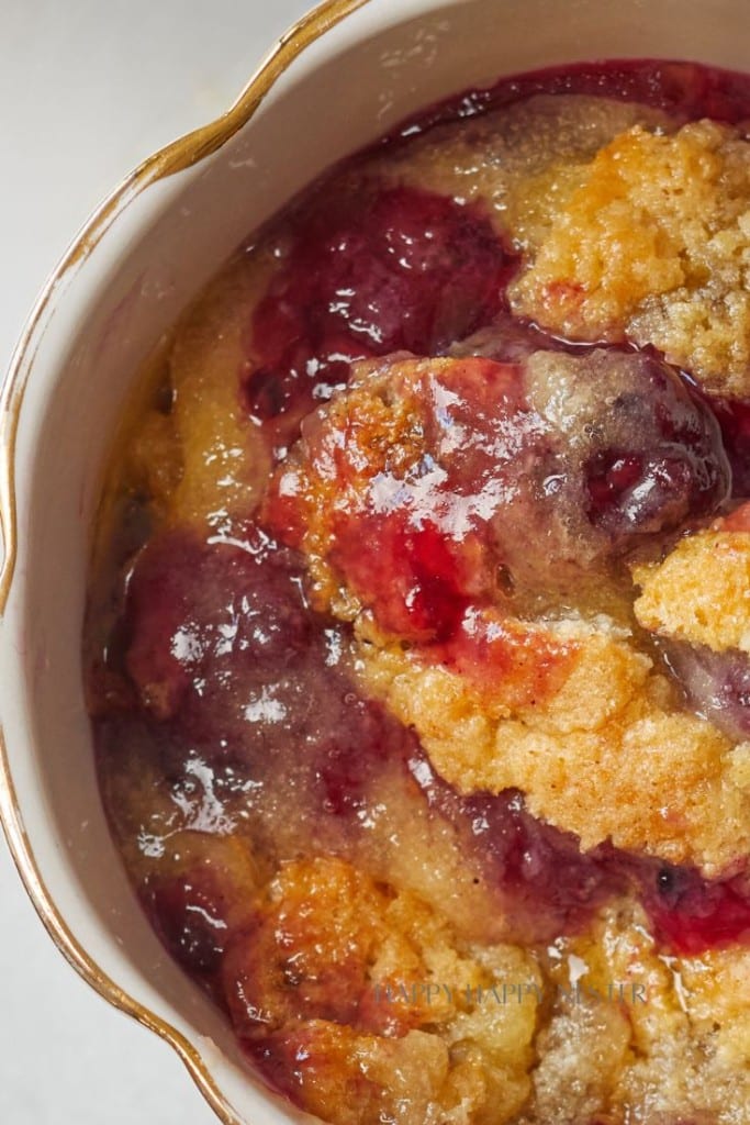 A close-up of a baked fruit cobbler in a white dish with a golden rim. The dessert has a golden brown crust and is topped with thick, bubbling fruit filling that appears to be a mix of red and purple berries, indicating a rich, sweet, and juicy texture.