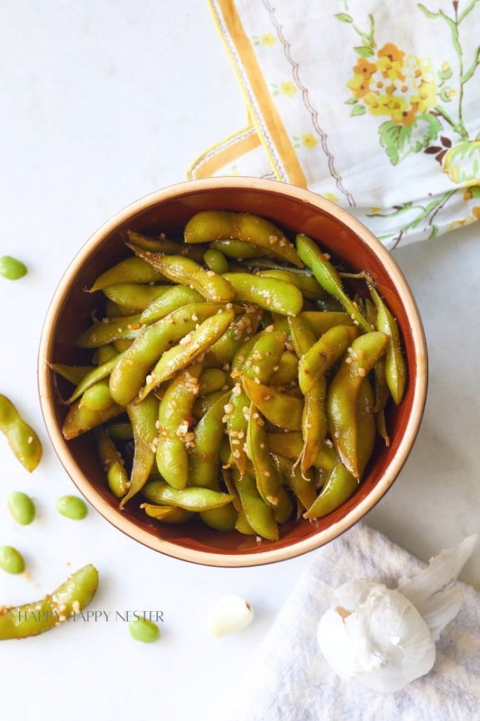 A bowl of seasoned edamame sprinkled with coarse salt, placed on a marble surface. A floral napkin is partly visible in the top right corner, and a garlic bulb and a cloth napkin are in the bottom right corner. Some edamame pods lie around the bowl.