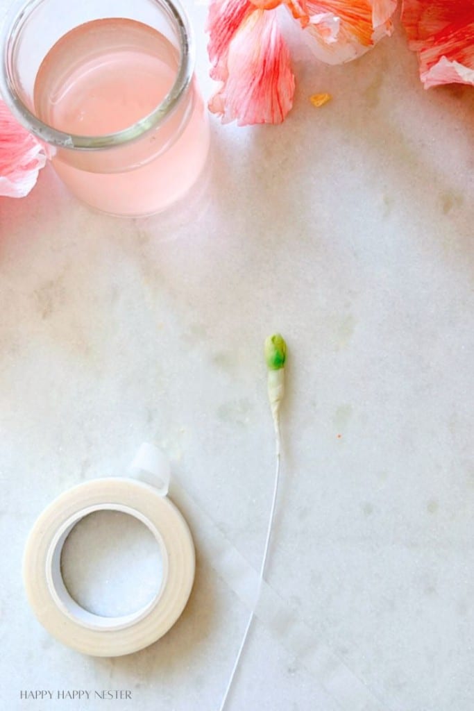 A white surface with a small glass jar filled with pink liquid, some pink flower petals, a roll of white tape, and a wire with a bud at the end.