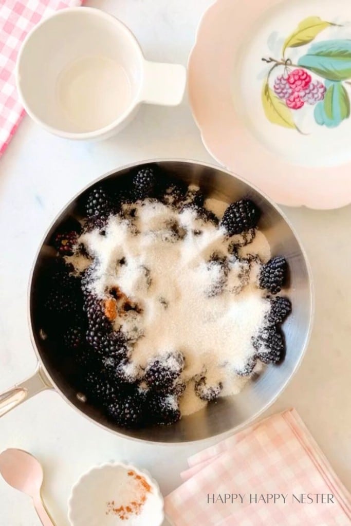 A saucepan filled with blackberries topped with granulated sugar rests on a white countertop. Surrounding the pan are a white teacup, a floral-patterned plate, a small bowl with ground spices, a pink spoon, and a pink checkered cloth.