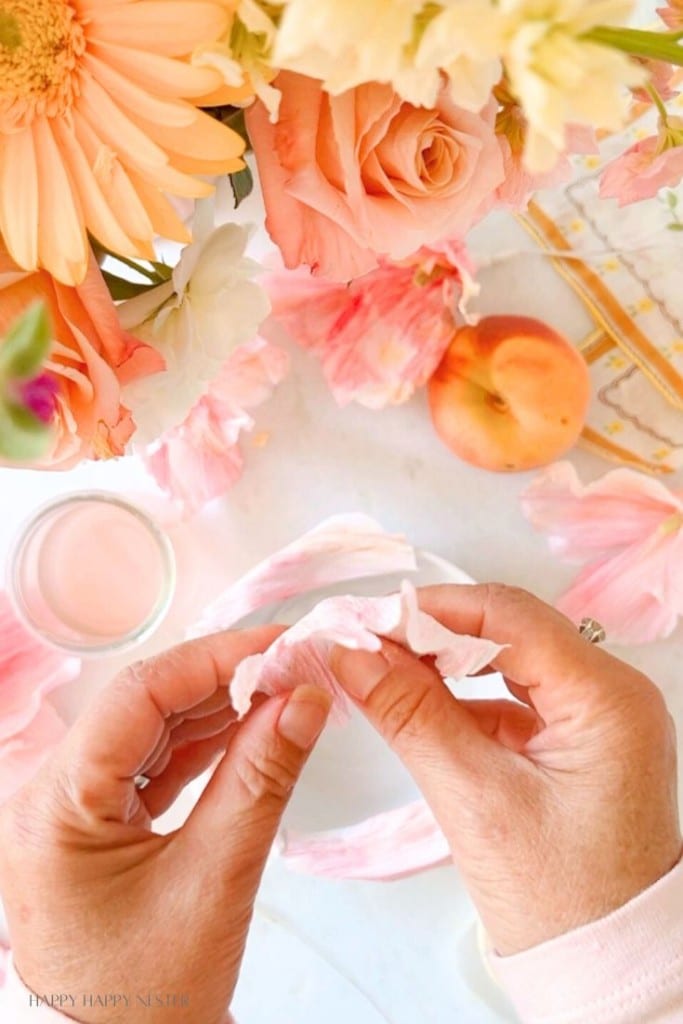 Two hands are seen crafting with pink crepe paper. Surrounding the hands are peach-colored flowers, an apricot, and a small bottle containing liquid. It's a delicate and colorful setup, suggesting a creative, floral-themed project.