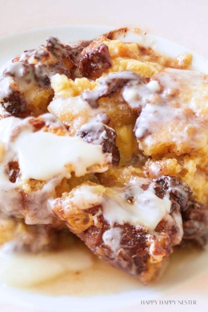 A close-up of a dessert featuring chunks of bread pudding drizzled with a generous amount of creamy glaze. The bread pudding has a golden-brown, caramelized texture with some darker, chocolatey sections.