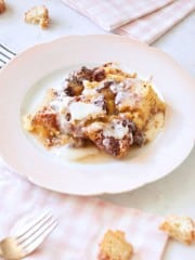 A white plate holds a serving of bread pudding drizzled with cream sauce. The table is adorned with a pink checkered cloth, a few scattered croutons, and two forks. The scene is bright and inviting, suggesting a homemade dessert setting.