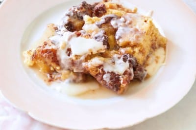 A white plate holds a serving of bread pudding drizzled with cream sauce. The table is adorned with a pink checkered cloth, a few scattered croutons, and two forks. The scene is bright and inviting, suggesting a homemade dessert setting.