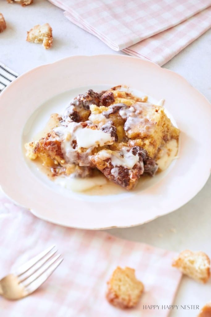 A white plate holds a serving of bread pudding drizzled with cream sauce. The table is adorned with a pink checkered cloth, a few scattered croutons, and two forks. The scene is bright and inviting, suggesting a homemade dessert setting.