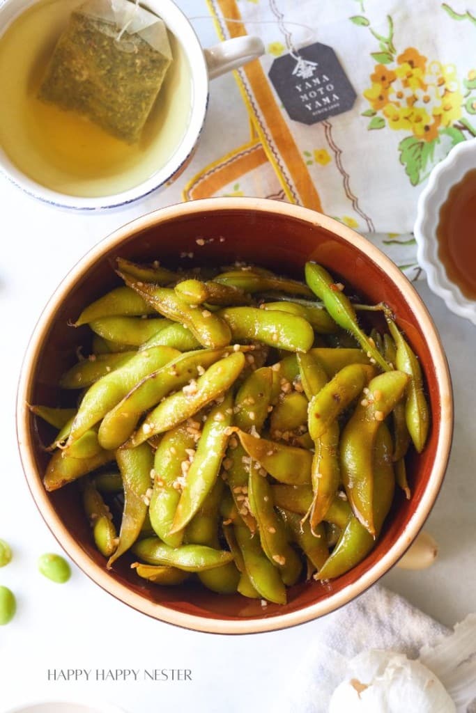 A bowl of seasoned edamame sprinkled with sesame seeds sits on a table adorned with a floral napkin. To the top left, there is a cup of tea with a tea bag. Around the bowl, some scattered edamame and a clove of garlic can be seen.