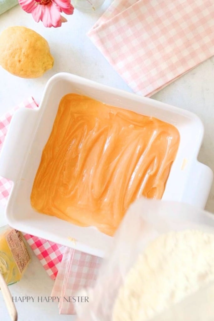 A white baking dish with an orange mixture spread inside is surrounded by a lemon, a pink flower, a checkered pink and white cloth, and baking ingredients such as flour and butter on a light-colored surface.