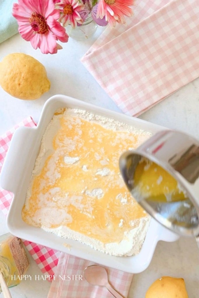 A baking dish filled with flour mixture is being covered with melted butter poured from a stainless steel pot. Lemons and pink flowers are nearby, along with a checkered cloth and a spoon. The setting is on a white surface.