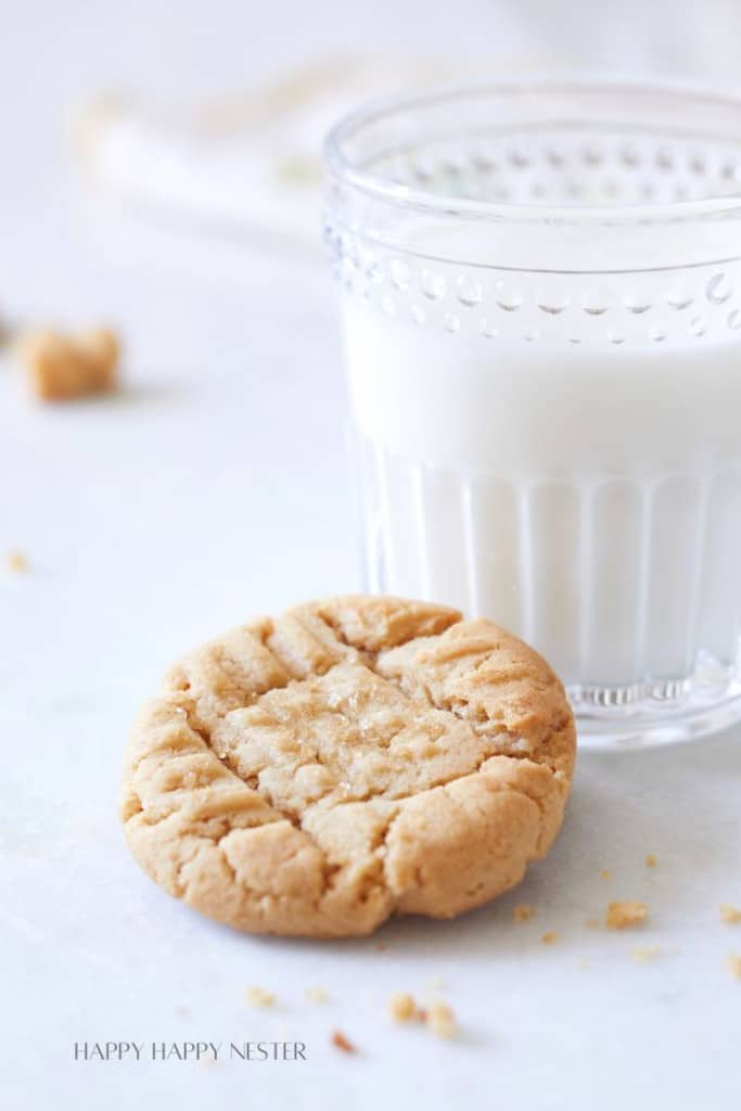 A close-up of a glass of milk and a peanut butter cookie on a white surface. The cookie has a crisscross pattern on top, with a few crumbs scattered nearby. The background is softly blurred, emphasizing the milk and cookie in the foreground.