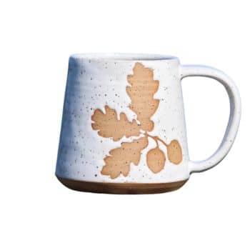 A white ceramic mug with a speckled design and a matte finish. The mug features a graphic of brown oak leaves and acorns on one side. The handle is white and the base of the mug is unglazed, showing the natural brown color of the clay.