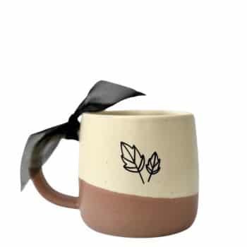 A ceramic mug with a two-tone design featuring a beige upper half and a light brown bottom half. The mug has a simple line drawing of two leaves on its side and a black ribbon tied around the handle.