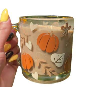 A hand with yellow and black painted nails holds a transparent mug filled with a creamy beverage. The mug is decorated with orange pumpkins, white hearts, leaves, and floral patterns, creating a festive autumnal theme.