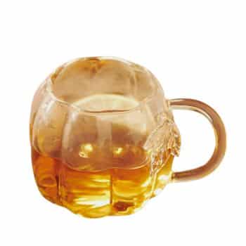 A small, transparent mug filled with a golden-brown liquid. The mug has a rounded, pumpkin-like shape with ridges and a short, sturdy handle. The liquid inside appears to be tea or another type of beverage.