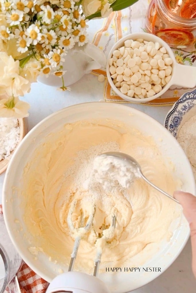 A hand holding a spoon adds flour to a mixing bowl filled with creamy batter. An electric mixer is partially submerged in the batter. Surrounding the bowl are white chocolate chips, flowers in vases, and other ingredients on a light-colored countertop.